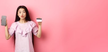 İnternetten alışveriş. Heyecanlı Asyalı kadın boş akıllı telefon ekranlı plastik kredi kartı gösteriyor. Reklam interneti, pembe arka planda duruyor..