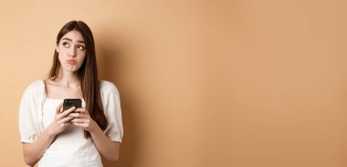 Kafası karışmış genç bir kadın cep telefonundan haberleri okuduktan sonra düşünüyor sol üst köşeye bakıyor tereddütlü, bej arka planda duruyor.