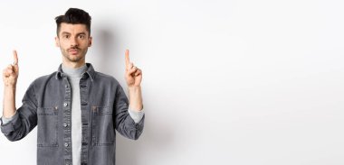 Bıyıklı, ciddi bir beyaz adam. Parmaklarını boş yere doğrultuyor ve beyaz arka planda kameraya bakıyor..