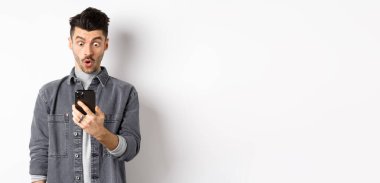 Adam akıllı telefon ekranına şaşırmış görünüyor, telefonda heyecan verici haberler alıyor, beyaz arka planda duruyor..