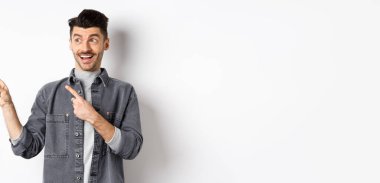 Yakışıklı, gülümseyen adam reklama bakıyor, bakıyor ve solu işaret ediyor, heyecanlı yüzüyle, beyaz arka planda duruyor..