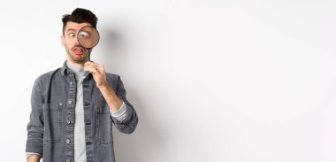 Komik genç adam gözlerini kısıyor, büyüteçle yüzlerini gösteriyor, beyaz arka planda duruyor..