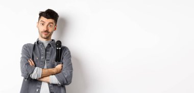 Ceketinin cebinde mikrofon tutan komik erkek şovmen ya da şarkıcı görüntüsü ve kollarını kavuşturup beyaz arka planda duran kameraya bakın..