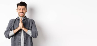 Lütfen bana yardım et. Gülümseyen sevimli adam namaste jestinde el ele tutuşuyor, iyilik istiyor, teşekkür ediyor, umut dolu bakıyor ve kameraya heyecanlı bakıyor, beyaz arka planda duruyor..