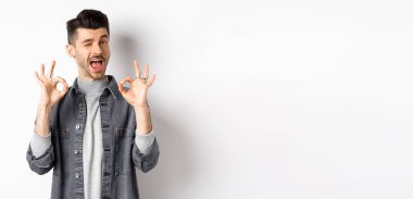 Her şey yolunda. Yakışıklı beyaz adam iyi işaretler gösteriyor ve gülümsüyor, iyi şeyi övmek için göz kırpıyor, mükemmel bir teklif sunuyor, beyaz arka planda duruyor..