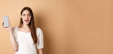 Akıllı telefon uygulaması gösteren neşeli stil kızı, boş telefon ekranını göster ve göz kırp, bej arka planda dur.