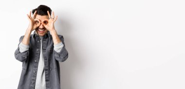 Gülümseyen komik adam, el dürbününden güzel bir teklif görüyor, mutlu ve heyecanlı görünüyor, reklamlara bakıyor, beyaz arka planda duruyor..