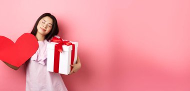 Sevgililer Günü konsepti. Romantik ve hassas Asyalı genç kız gülümsüyor, gözlerini kapatıyor ve sevgilisinin hediyeleriyle hayaller kuruyor, hediyesini tutuyor ve büyük kırmızı kalp kesimi, pembe arka plan.