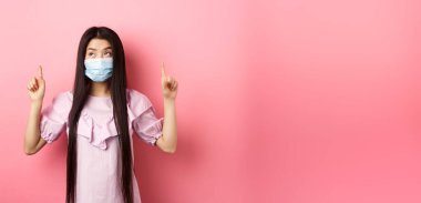 Coronavirus, karantina ve yaşam tarzı konsepti. Tıbbi maskeli sevimli Koreli kız parmaklarını kaldırmış, en iyi reklamlara bakıyor, pembe arka planda duruyor..