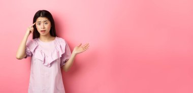 Delirdin mi sen? Şok olmuş Asyalı bir kadın baş tapınağı işaret ediyor ve birinin aptalca hareketlerinden şikayet ediyor, insanları azarlıyor, pembe arka plana karşı duruyor..