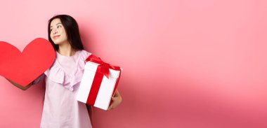 Sevgililer Günü konsepti. Aşk hediyeleri tutan romantik kız, büyük kırmızı kalp kartı ve hediye kutusu, rüya gibi bakışlar, aşık olmak, şefkatle gülümsemek, pembe arka plan.
