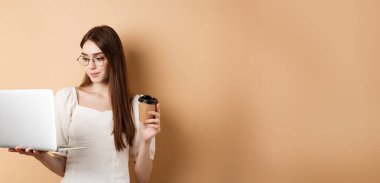 Dizüstü bilgisayarda çalışan ve kahve içen kadın portresi. Uzaktan çalışmalar için bilgisayar kullanan kız öğrenci, bej arka planda duruyor.