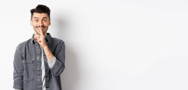 Mutlu adam kameraya şşşş yapıyor, parmakları dudaklarında gülümsüyor, sessiz kalmak istiyor, büyük bir sır veriyor ya da sürpriz yapıyor, beyaz arka planda duruyor..