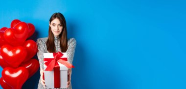 Sevgililer Günü. Romantik ve tatlı bir kız hediye kutusunu sevgilisine veriyor ve gülümsüyor, kalp balonlarının yanında duruyor, mavi arka plan..