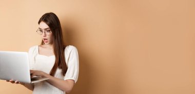 Endişeli kız dizüstü bilgisayara bakar, odaklanmış yüzüyle bilgisayarda yazar. İnternette çalışan serbest çalışan, bej arka planda duran bir kadın..