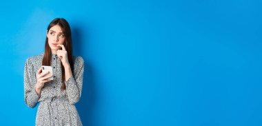 Düşünceli şirin kız mesajda nasıl cevap vereceğini düşünüyor, düşünceli ve akıllı telefonunu tutuyor, mavi arka planda elbise giyiyor..