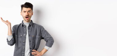 Kafası karışık ve sinirli adam elini kaldırıp şikayet ediyor, ne olduğunu soruyor, anlayamıyorum, beyaz arka planda duruyor..