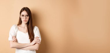 Kızgın ve gücenmiş gözlüklü bir kız. Kaşlarını çatmış, kollarını göğsüne dayamış ve somurtuyor. Bej arka planda savunma yapıyor..