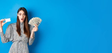 Heyecanlı şanslı kız plastik kredi kartı ve para gösteriyor, hayret içinde, pahalı bir şey alıyor, mavi arka planda duruyor..