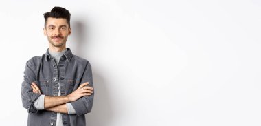 Kendine güveni tam yakışıklı bir adam göğsünde çapraz kollar, gülümsüyor, beyaz arka planda profesyonel duruyor..