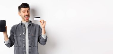 Neşeli yakışıklı adam iyi bir anlaşma yapıyor, boş akıllı telefon ekranı ve plastik kredi kartı, sana göz kırpıyor ve gülümsüyor, öneri sunuyor, beyaz arka plan..