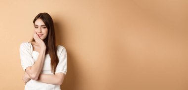 Romantik genç bir kadın yüzüne dokunuyor ve kameraya dalgın dalgın bakıyor, bej arka planda rüya gibi duruyor..