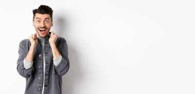 Heyecanlı genç adam mutluluktan atlıyor, şaşırmış bir şekilde gülümsüyor ve el ele tutuşuyor, beyaz arka planda duruyor..