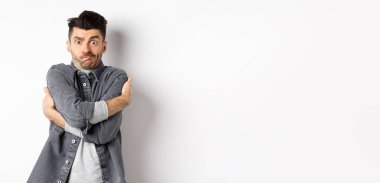 Komik genç adam kendini kucaklıyor, yüzünü buruşturuyor, utanmış ve teşhir edilmiş hissediyor, vücudunu insanlardan gizliyor, beyaz arka plan..