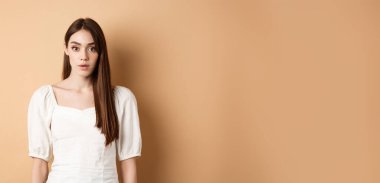 Şok olmuş ve ürkmüş genç bir kadın huşu içinde donup kalıyor, ağzı açık kalıyor ve kameraya hayret içinde bakıyor, bej arka planda giyinmiş duruyor..