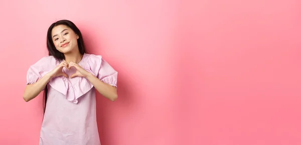 Valentines day concept. Beautiful korean teenager in dress showing heart gesture and smiling with admiration and affection, falling in love, feel romantic, pink background.