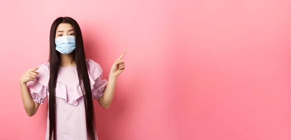 Healthy people and covid-19 pandemic concept. Bored asian woman in medical mask pointing right, showing logo, standing unamused on pink background.