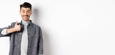 Heyecanlı yakışıklı adam baş parmağını gösteriyor ve gülümsüyor, iyi bir şey gibi, harika seçimi övüyor, tatmin olmuş ve mutlu görünüyor, beyaz arka planda duruyor..