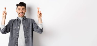 İyimser genç erkek, hayallerinin gerçekleşmesini umuyor, parmaklarını çapraz tutuyor, gülümsüyor ve iyi haberler bekliyor, beyaz arka planda duruyor..