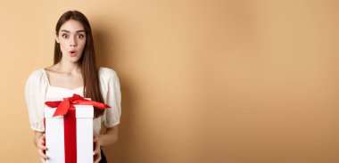 Sevgililer Günü 'nde hediye alan şaşırmış bir kızın görüntüsü. İnançsızlıkla bakıyor ve bej arka planda duruyor..