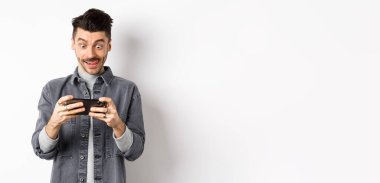 Heyecanlı adam cep telefonuyla video oyunu oynuyor odaklanmış yüzü, akıllı telefon ekranını dinliyor, cep telefonunu yatay olarak tutuyor, beyaz arka plan.