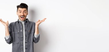 Heyecanlı genç adam sevinçten havalara uçuyor, ellerini yana açıyor ve mutlu görünüyor, şaşkın şaşkın gülüyor, beyaz arka planda duruyor..