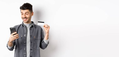 İnternetten alışveriş konsepti. Gülümseyen adam cep telefonuyla ödeme yapıyor, plastik kredi kartı gösteriyor, internetten alıyor, beyaz arka plan.