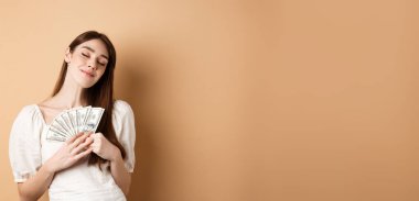 Zarif bir kız, mutlu bir gülümsemeyle ve kapalı gözlerle parayı kucaklıyor, dolar banknotları gibi, bej arka planda duruyor..