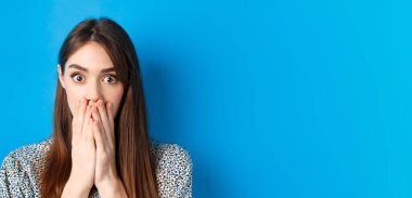 İnsanların duygu konsepti. Şok olmuş genç bir kadın yüzü, elleri ve soluk soluğa, ürkmüş görünüyor, mavi arka planda duruyor..