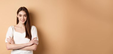 Göğsünde çapraz kollar olan, gülümseyen samimi kız, profesyonel gibi görünüyor, bej arka planda kendine güveniyor..