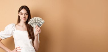 Dolarları gösteren, para kazanan ve bej arka planda nakit parayla duran kendine güvenen, güler yüzlü bir kadın..