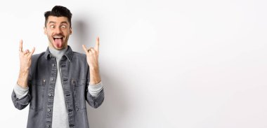 Dilini ve kaya boynuzlarını gösteren komik genç adam, eğlenmek için gözlerini kısarak bakan, parti ya da konserin tadını çıkaran, beyaz arka planda duran.
