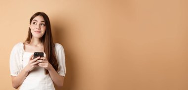 Telefonla düşünen romantik bir kız, kenara bakıyor ve gülümsüyor, akıllı telefonda flört uygulaması kullanıyor, bej arka planda duruyor..