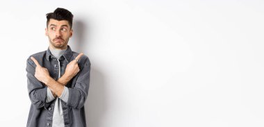 Kararsız yakışıklı adam parmaklarını yana çeviriyor, yana bakıyor ve seçimler yapıyor, iki varyantı gösteriyor, beyaz arka planda kararsız duruyor..