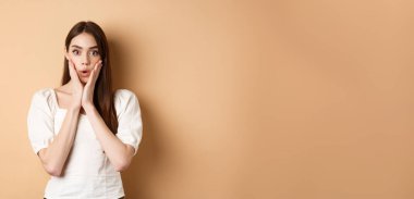 İnanılmaz. Etkilenmiş genç bir kadın nefes nefese kalmış ve haberlere hayran kalmış, kameraya bakıyor, huşu içinde yüzüne dokunuyor, bej arka planda duruyor..