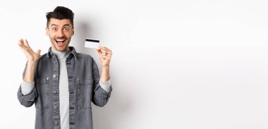 Heyecanlı adam sevinçten çığlık atıyor ve plastik kredi kartı gösteriyor, alışveriş yaparken seviniyor, beyaz arka planda duruyor..