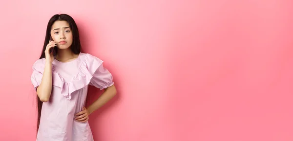 Sad asian girl calling someone, holding phone and talking, standing upset against pink background.