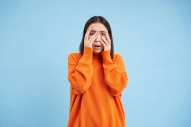 Korkak Asyalı kadın gözlerini kapar ve elleri üzerinden korkunç ve rahatsız edici bir şekilde mavi arka planda dikilirken şok geçirir..