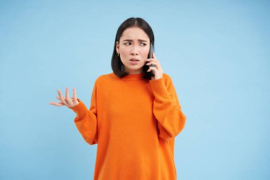 Kafası karışmış bir kadın cep telefonunu kaldırıyor, kafası karışmış bir şekilde akıllı telefondan konuşmalar dinliyor, omuz silkiyor, mavi arka planda dikiliyor..