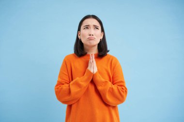 Tanrı 'ya dua et. Sefil Asyalı kız dua ederken el ele tutuşur, dua eder, kafasını kaldırır ve mucizeyi bekler, mavi arka planda durur..